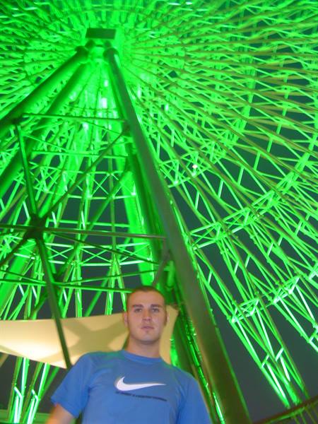 Man in Nike t-shirt standing beneath a green-lit Ferris wheel at night