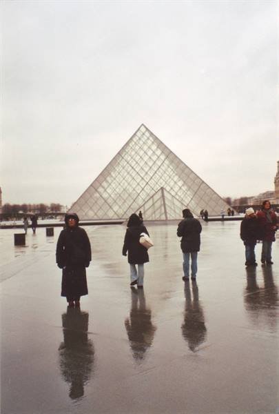 People in coats walk across wet pavement in front of the Louvre glass pyramid