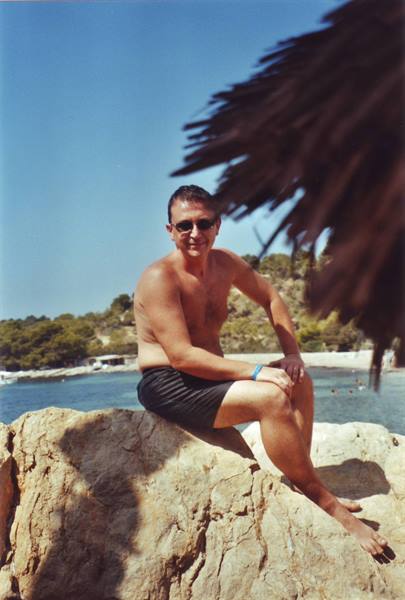 Man in black swimsuit and sunglasses sitting on a rock by the bay