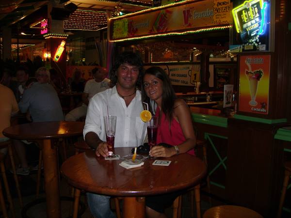 Smiling couple with two cocktails at a wooden table in a busy bar with neon signs