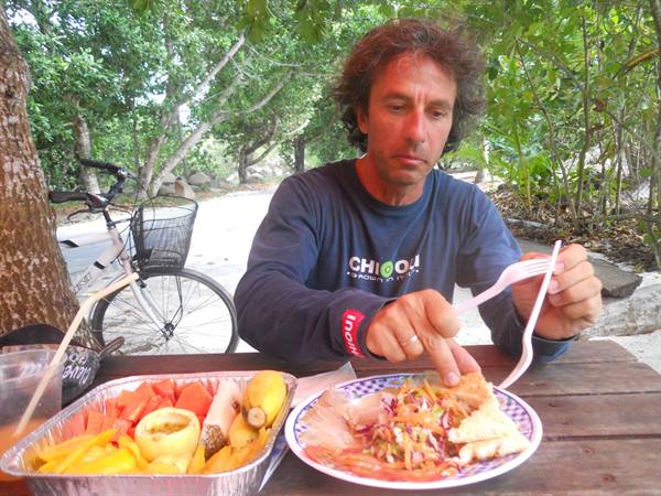 Man eating fish with fork at wooden table, bicycle parked among trees