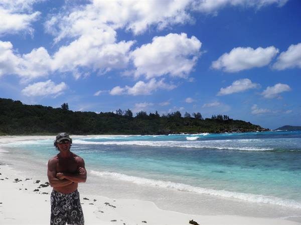 Man with crossed arms on white sand beach, turquoise sea and green trees