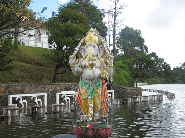 Painted Ganesha statue on lotus, standing in lake water among trees and white chapel