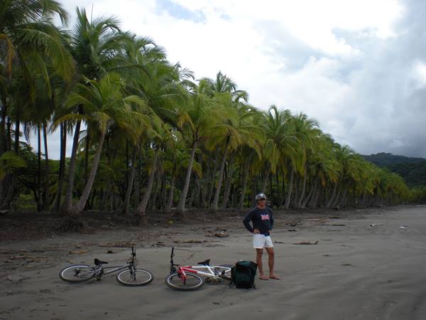 Cyclist standing on the beach beside two bicycles under coconut palms
