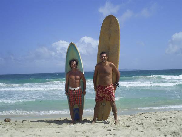 Two surfers in shorts holding their boards on the beach, waves and blue sky