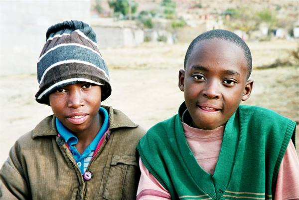 Two boys smile at the camera, one in a striped beanie, village behind