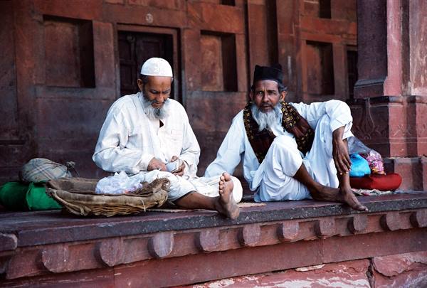 Two bearded elders seated on red sandstone steps with baskets and bags