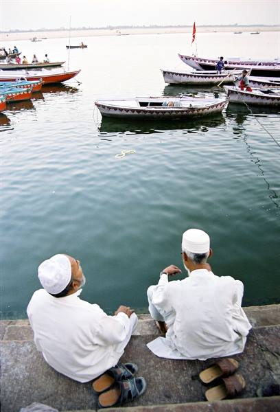 Two men in white sit on stone ghat steps watching boats on the river