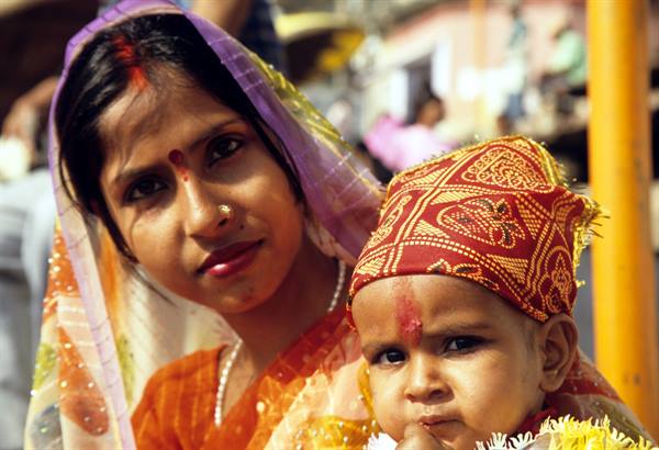 Veiled Indian woman with child; red tikkas on foreheads, orange sari, brocade cap