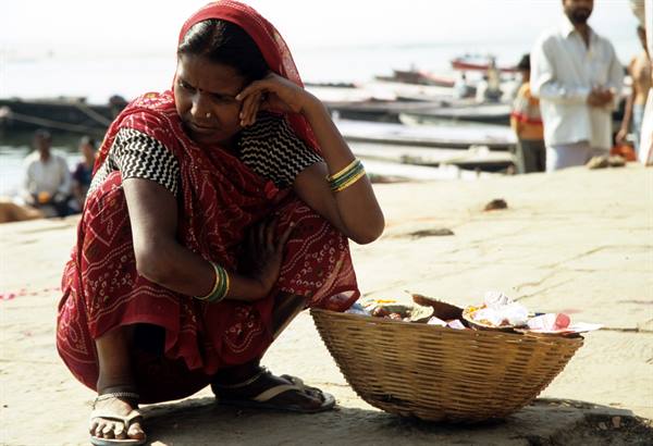 Woman in red sari crouching beside a basket, river ghat with boats, Varanasi