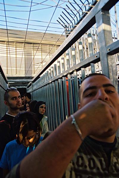 People queuing behind tall spiked metal fences under bright sun