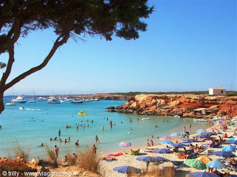 Crowded beach with colorful umbrellas, reddish rocks and boats, maybe Cala Saona