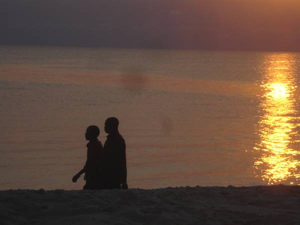 Two silhouettes walk on the sand, the sun reflects golden on the water