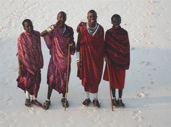 Four young Maasai men in red shukas with wooden staffs on white sand