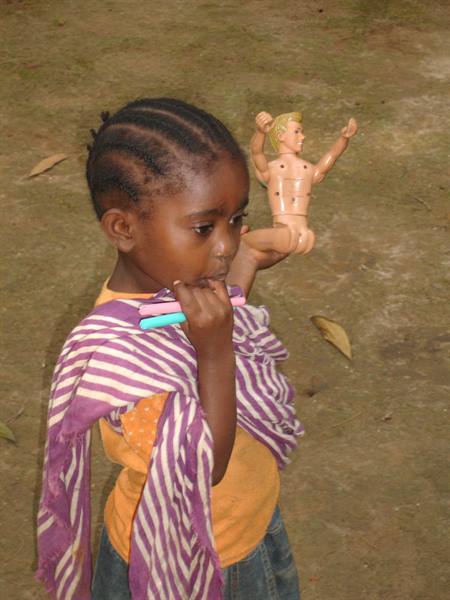 Girl in purple striped shawl holds a blond doll on bare dirt ground