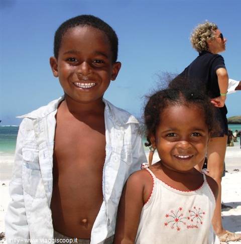 Two smiling children on white sand beach, blonde woman behind them