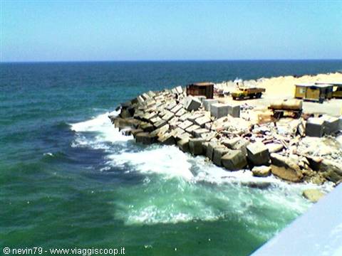 Waves crash on concrete breakwater blocks, trucks and containers under the sun