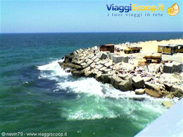 Waves crash on concrete breakwater blocks, trucks and containers under the sun