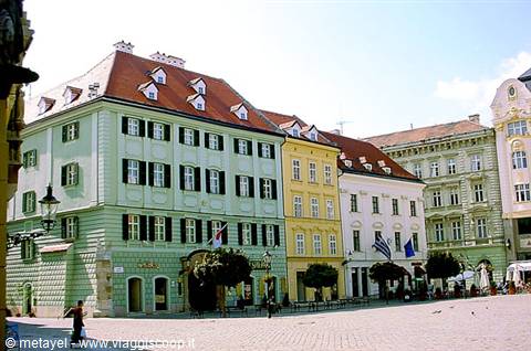 Pedestrian square with pastel buildings and red roofs, sunlight, a walker passes