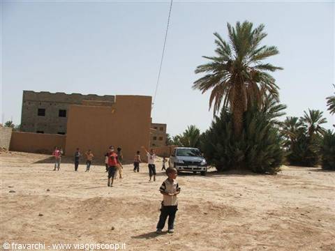 Child on sandy ground, mud-brick houses, palm trees and a parked car