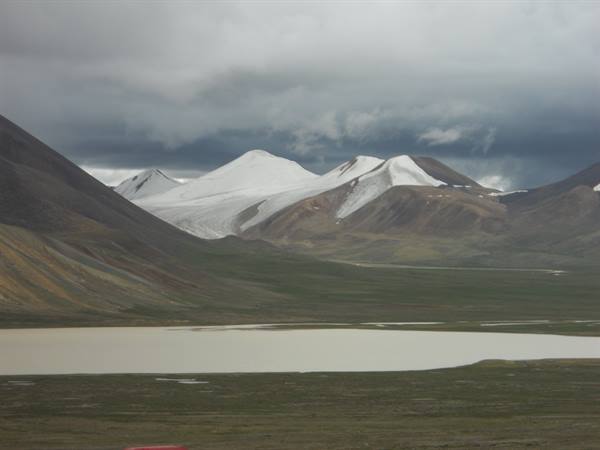 Glacial lake below snow-capped mountains under dark clouds, green valley