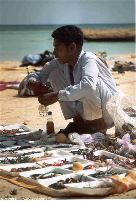 Young vendor crouching on the beach beside trays of colorful handmade jewelry