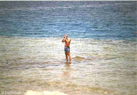 Man in swim trunks standing in shallow sea water taking a photo