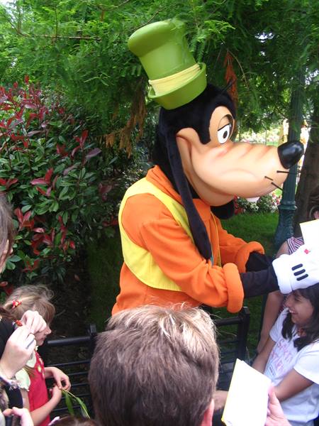 Goofy mascot greets children among green trees at Disney park