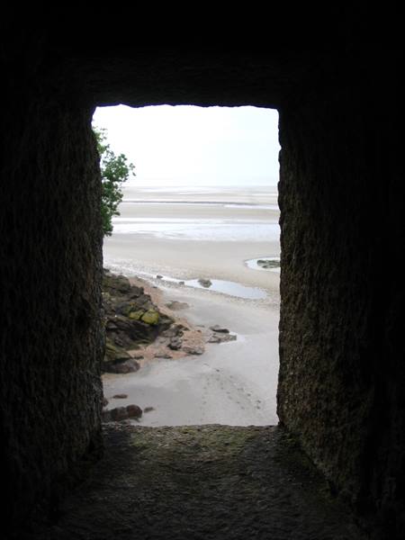 Through a dark stone window, a sandy bay with seaweed-covered rocks