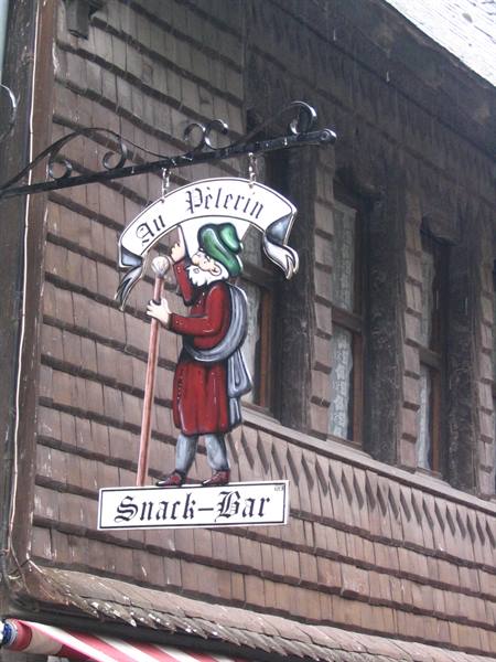 Metal sign of the Au Pèlerin pub with painted pilgrim, on wooden shingle facade