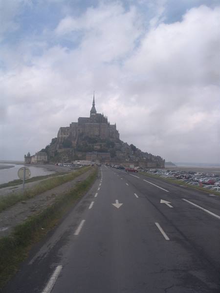 Road with white arrows leading to Mont-Saint-Michel abbey under cloudy sky