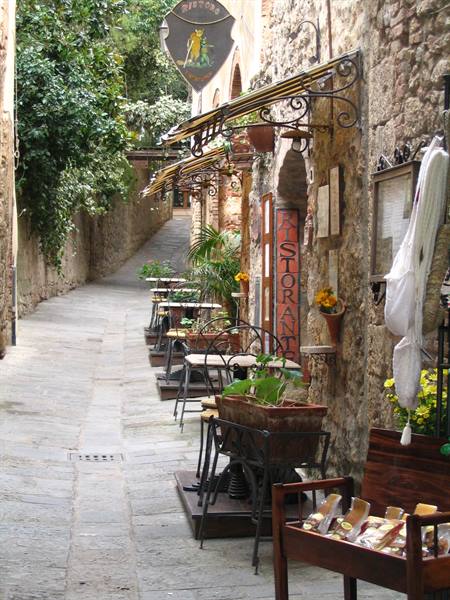 Cobbled alley with restaurant tables under wrought-iron awnings, stone walls
