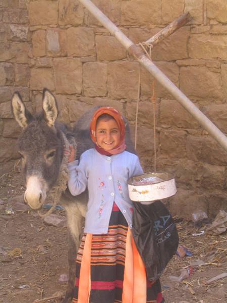 Girl in orange headscarf pets a donkey beside a stone wall