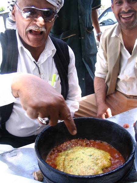 Man in turban points at omelette in black pan, friend laughing beside