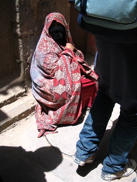 Veiled woman wrapped in red patterned shawl sitting on stone steps in sunlight
