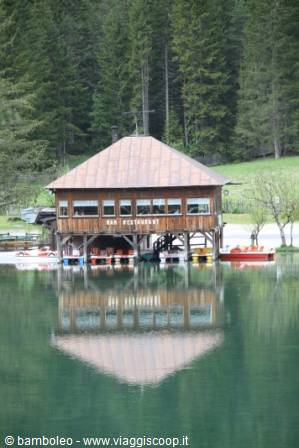 Wooden boathouse reflected in green lake water, rowing boats moored below, pines behind