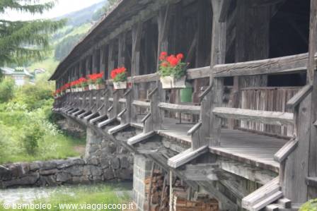 Covered wooden bridge with red geraniums on the railings, stream and mountains