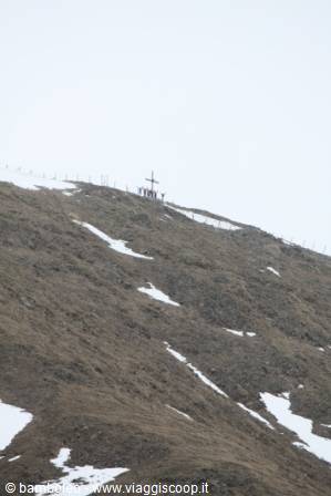 A metal cross on a grassy slope with snow patches, people gathered around