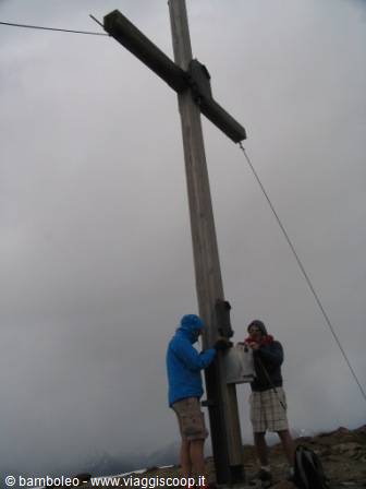 Two hikers beneath a wooden summit cross, grey foggy sky above