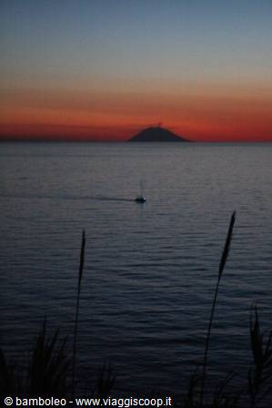 Stromboli volcano silhouetted at dusk, a small boat crosses the dark sea