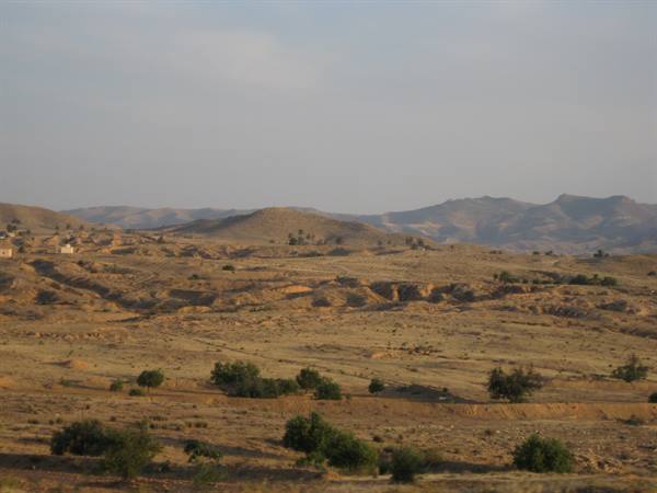 Arid hills and scattered shrubs under hazy sky, distant houses on the left