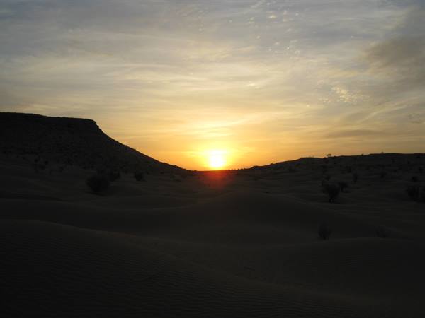 Low sun between sand dunes, dark rocky hill and scattered shrubs