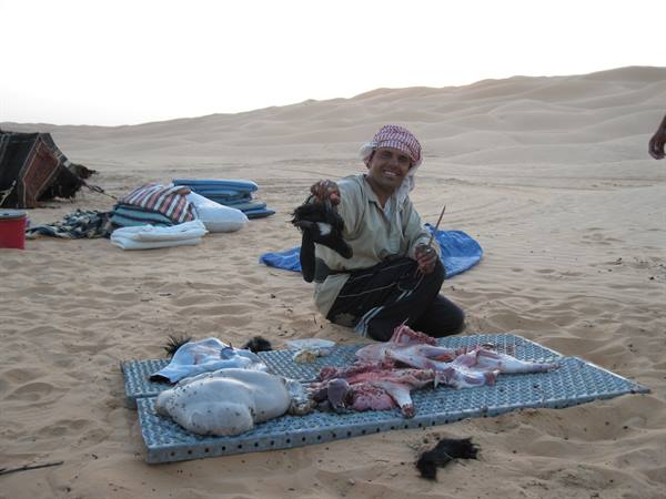 Man crouching butchers a carcass on a metal mat in sandy desert