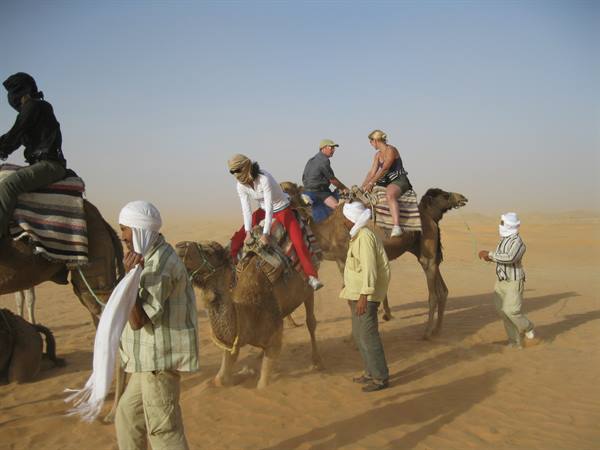 Tourists riding camels on sand, guide in white turban beside them