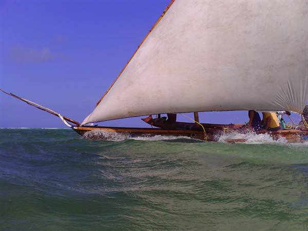 Wooden dhow with white sail cuts through green waves, men on board