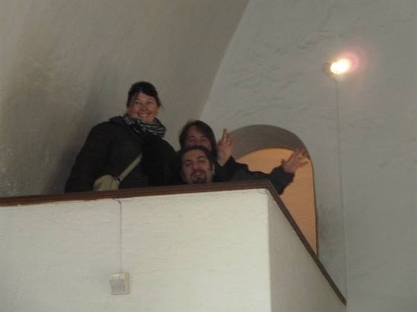 Three visitors waving from a white indoor balcony under warm light