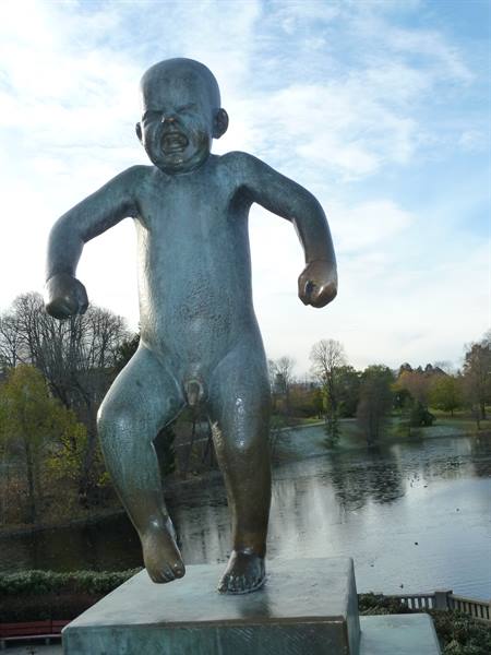Bronze statue of a crying baby on a pedestal near an autumn lake, daylight
