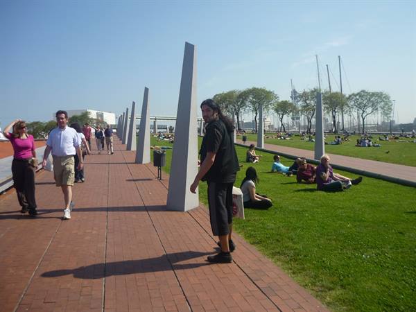 Brick waterfront promenade with concrete obelisks, people strolling and resting on grass