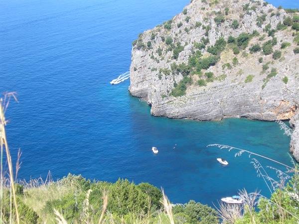 Rocky cliffs over turquoise cove, anchored boats, grass in foreground