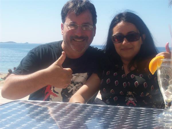 Smiling couple at a bar table, thumbs up, sea and beach behind